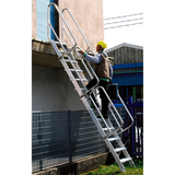 Ouvrier sur chantier utilisant un escabeau pliable en aluminium réglable, fixé à une dalle béton avec garde-corps intégral pour un accès sécurisé en hauteur.